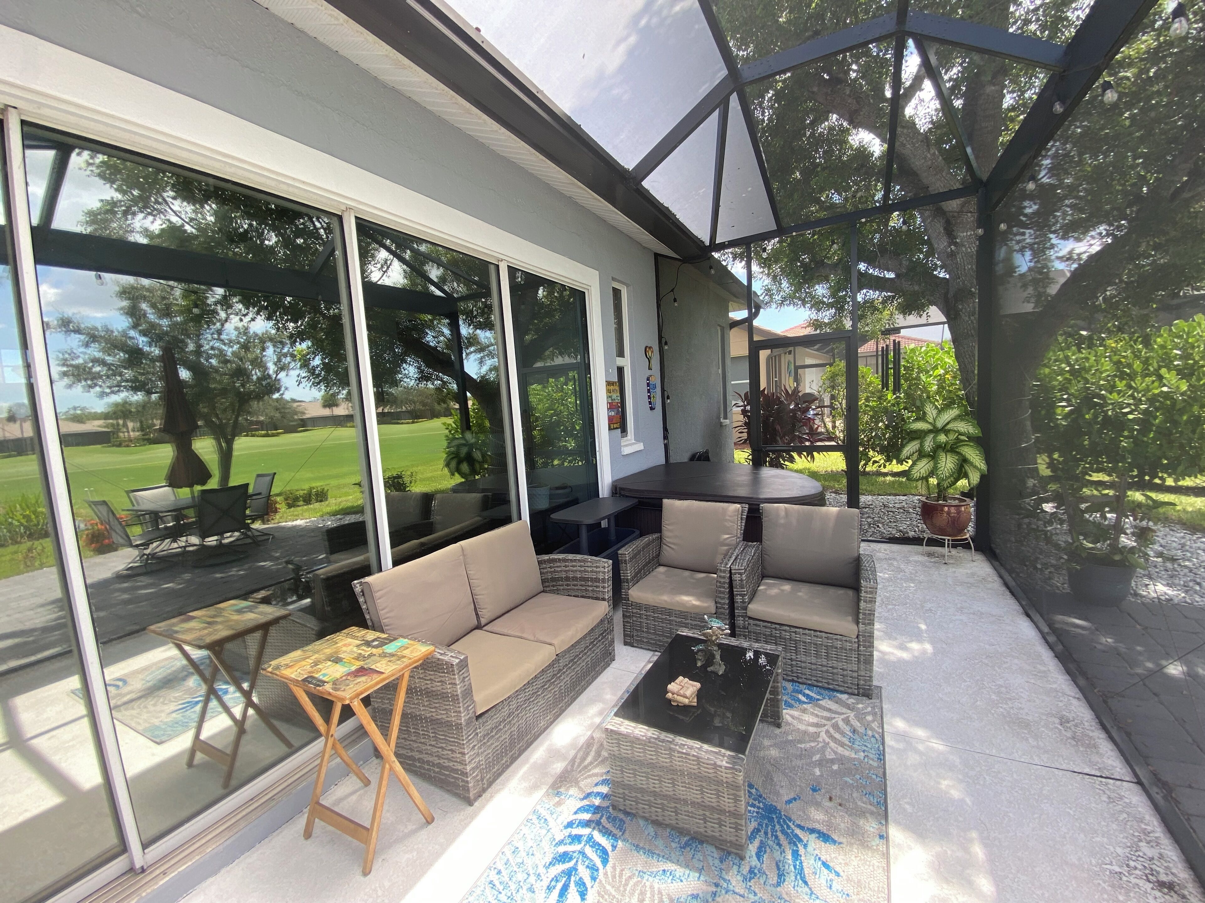 A covered patio area with outdoor furniture, including a sofa and a table, and a view of a green landscape through large windows.