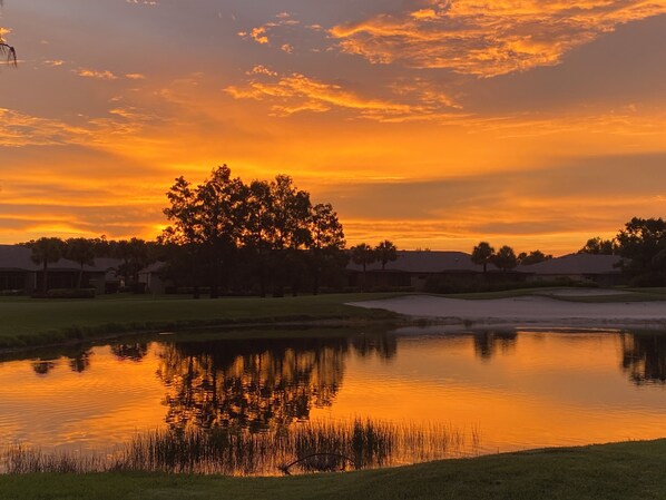Un atardecer tranquilo sobre un campo de golf con un estanque que refleja el cielo, rodeado de árboles y casas.