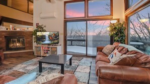 Ein Wohnzimmer mit Kamin, Fernseher, Deckenventilator und großen Fenstern mit Blick zum Himmel.