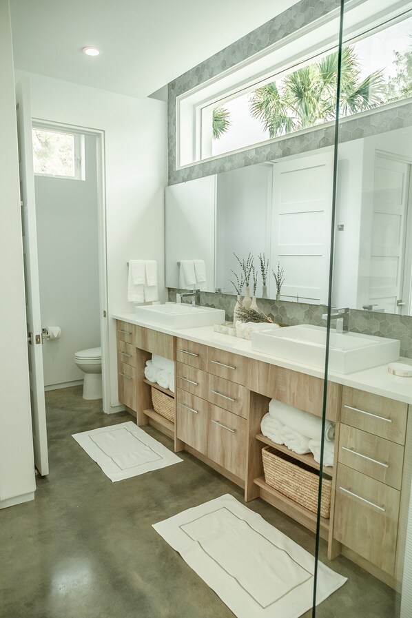 A modern bathroom with a large mirror, two sinks, wooden cabinets, and a view of palm trees through the window. - Location, comfort, and 5 beach accesses to more than one mile of deeded beach! (Seagrove Beach)