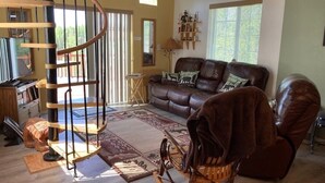 A spacious living room with a brown leather sofa, a rocking chair, a spiral staircase, and a bookshelf.