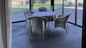 Un espacio de estar al aire libre techado con mesa y sillas, ventanas amplias y vistas a un jardín ajardinado.