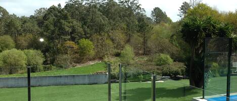 A view from a balcony with artificial grass, a pool, and a forested hillside.