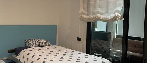 A bedroom with two beds, a large window with sheer curtains, and a view of a balcony.