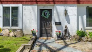 Une maison à l’extérieur blanc, avec une porte en bois foncé et une guirlande accrochée à la porte. De part et d’autre de la porte, on trouve des plantes en pot ornées de fleurs rouges.