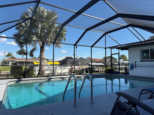 A pool with a clear blue water, surrounded by a covered area with a metal frame and glass panels, palm trees, and a view of houses in the background. - Waterfront Property in the heart of beautiful Cape Coral, FL (Cape Coral)