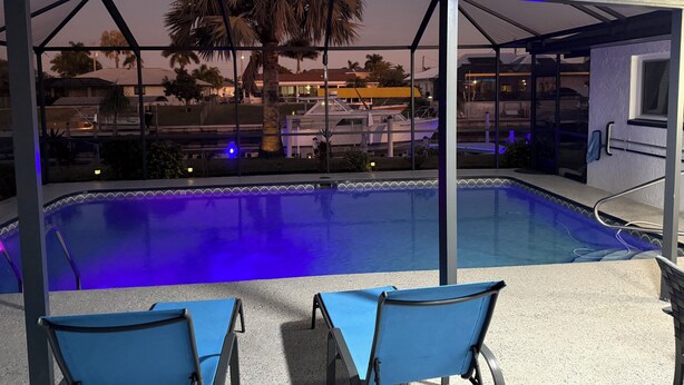 En pool med blått ljus, två blå stolar, ett palmträd och utsikt över en byggnad med en båt förtöjd utanför.