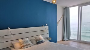 Een slaapkamer met een groot bed, een hoofdeinde met ingebouwde verlichting, een nachtkastje en uitzicht op zee door een groot raam.