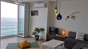 Een moderne woonkamer met een televisie, bank en een eettafel. Er is een groot raam met uitzicht op de oceaan.