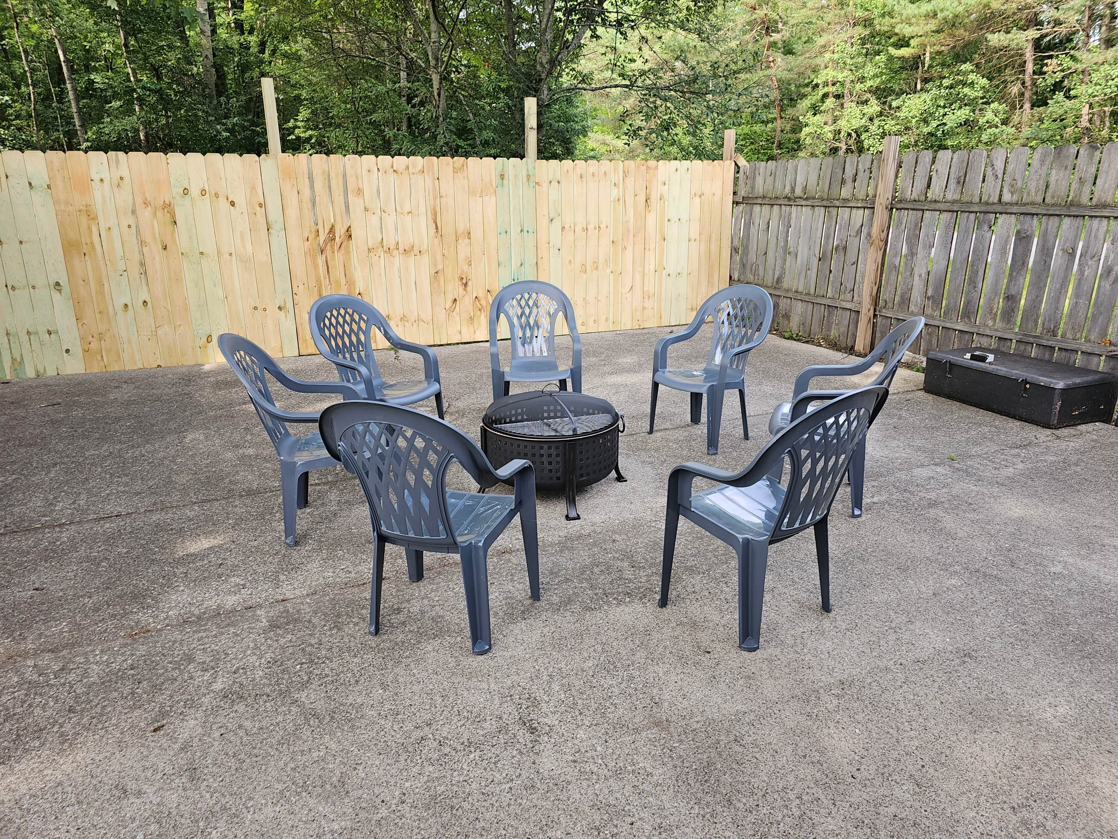 Un jardin avec une terrasse en gravier, une clôture en bois et six chaises en plastique bleues disposées autour d’un barbecue noir.