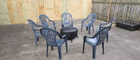 Un jardin avec une terrasse en gravier, une clôture en bois et six chaises en plastique bleues disposées autour d’un barbecue noir.