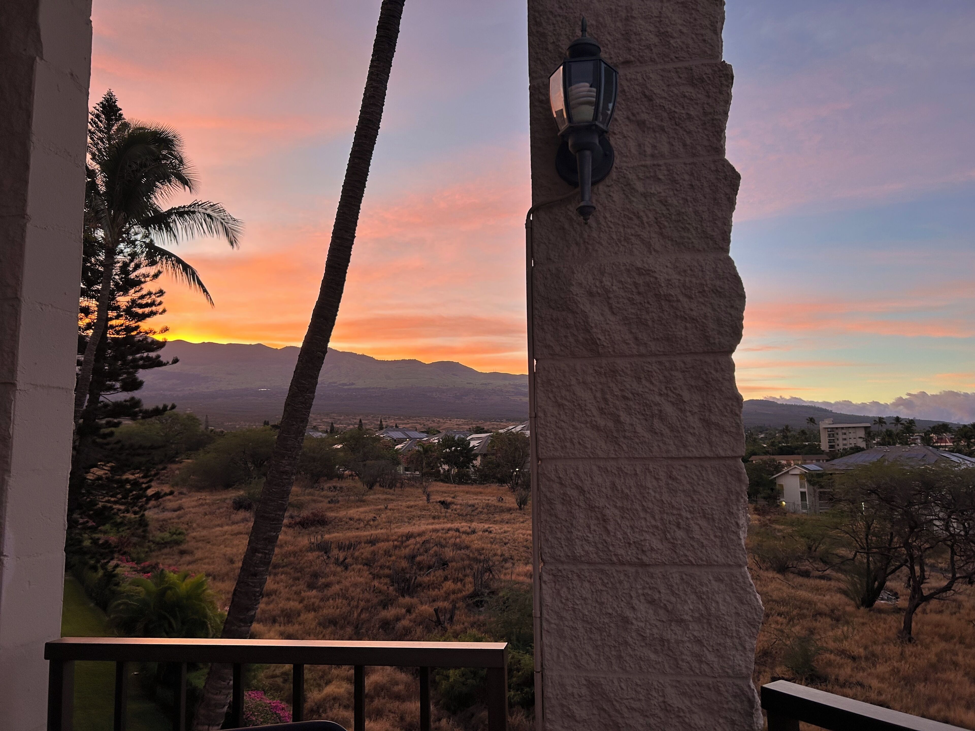 Un balcon offrant une vue sur un coucher de soleil se déclinant sur un paysage de montagnes et d’un palmier.