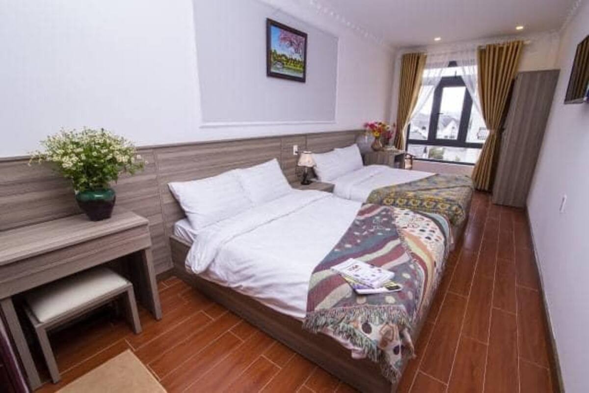 A hotel room with two beds, a desk, a chair, a window with curtains, and a potted plant.