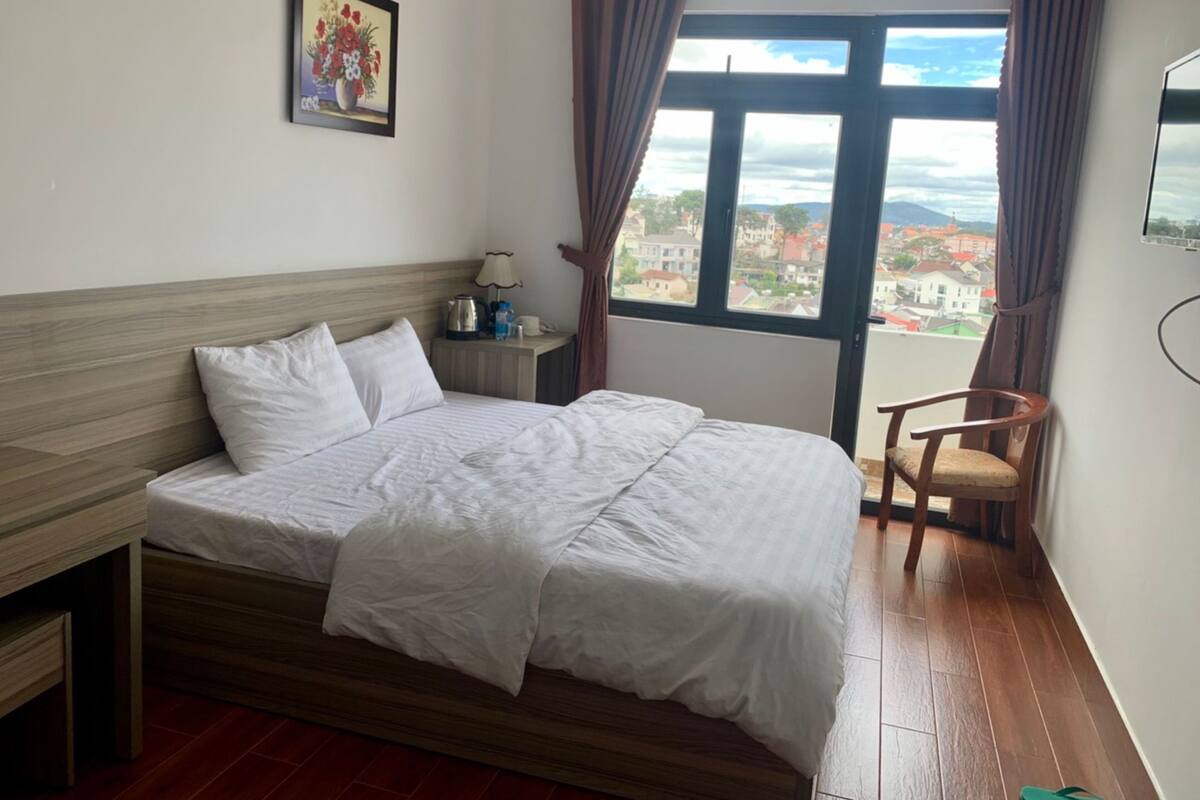 A hotel room with a bed, a nightstand, a chair, a window with a view, and a painting on the wall.