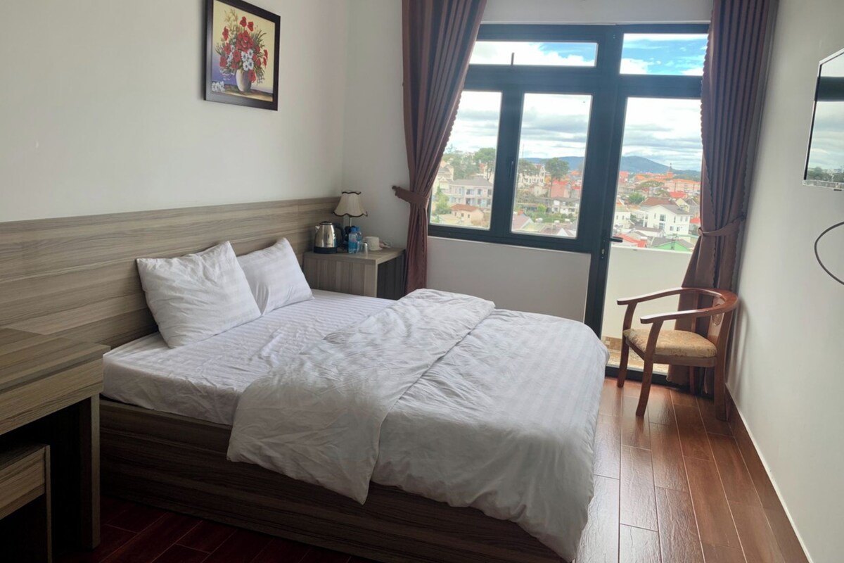 A hotel room with a bed, a nightstand, a chair, a window with a view, and a painting on the wall.