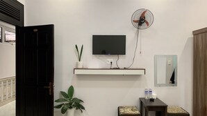 Una habitación con televisión montada en la pared, un ventilador de pie, una mesita con un banco, una planta en maceta y un espejo.