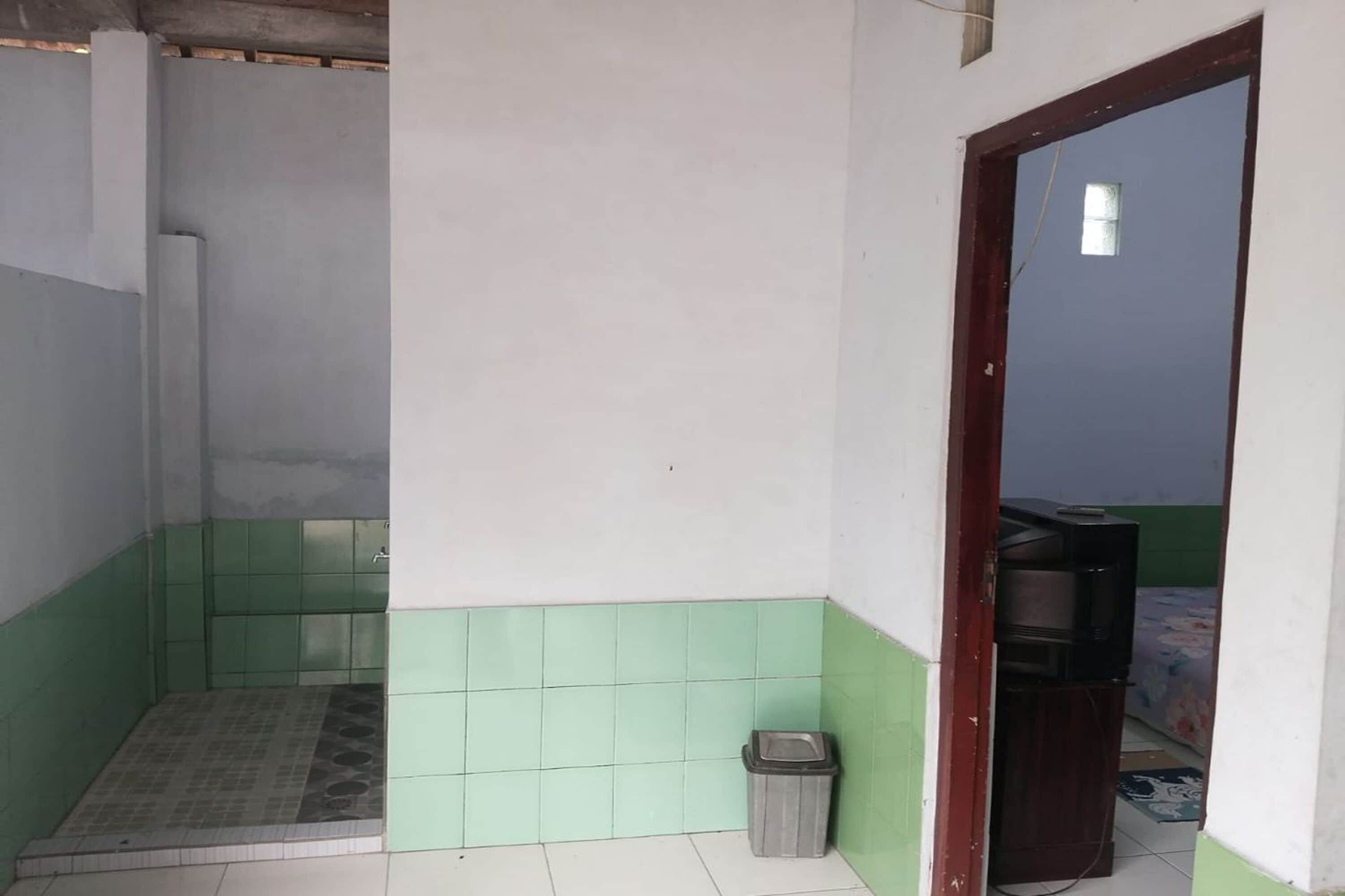 A small, tiled bathroom with a shower area, a trash bin, and a wooden door leading to another room.