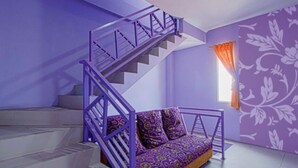 Una habitación con sofá púrpura, escalera con barandilla púrpura y ventana con cortinas anaranjadas.