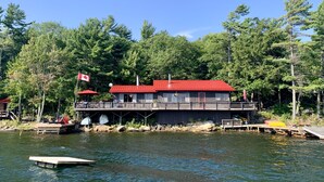 A lakeside cabin with a red roof, dock, and a Canadian flag. - WestWind Georgian Bay Muskoka Cottage Retreat Private Access (Sleeps Max 8) (MacTier)