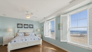 Una camera da letto con un letto, comodini, una ventola a soffitto e vista sulla spiaggia attraverso una finestra.