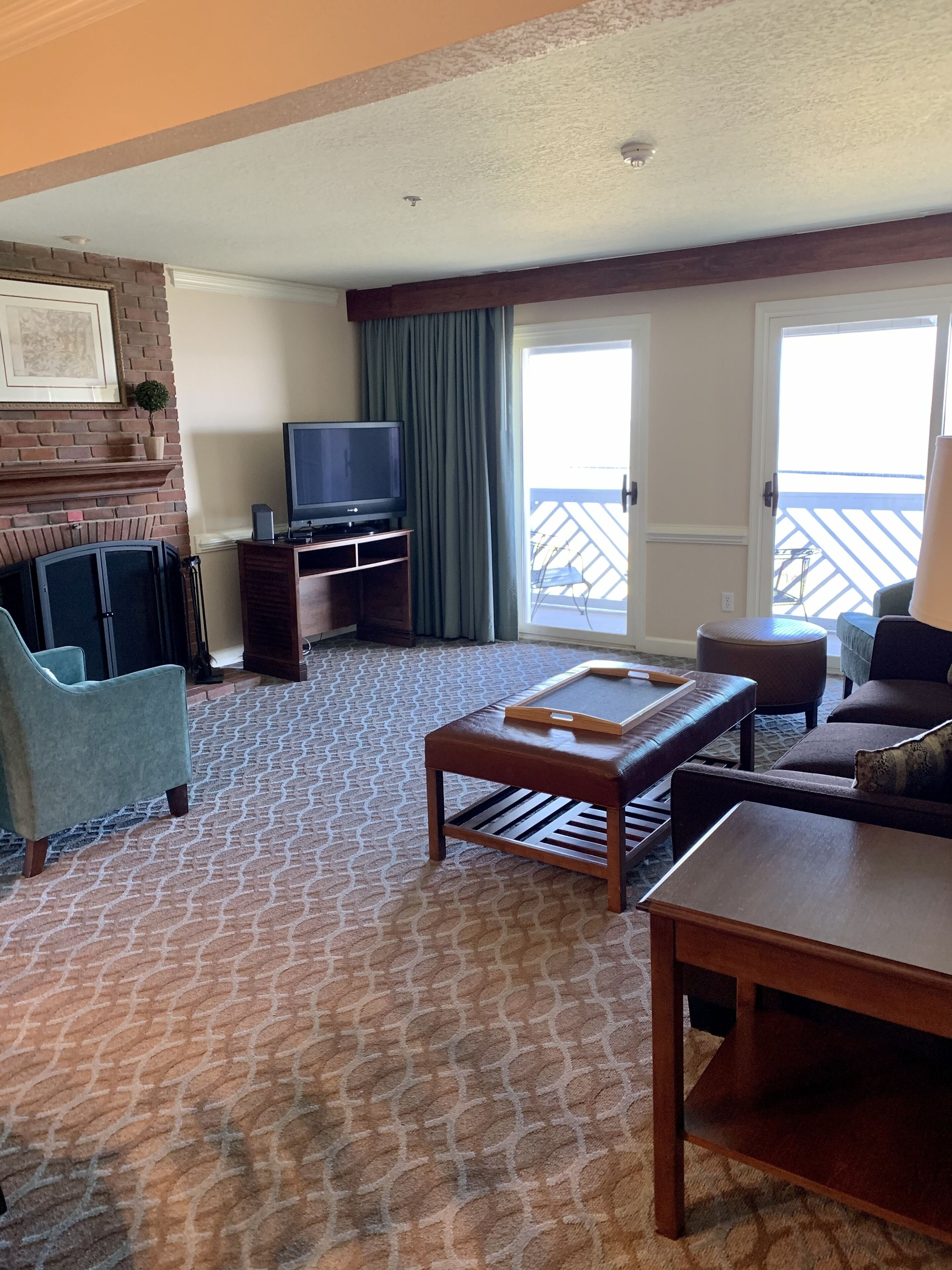 A hotel room with a patterned carpet, a television on a wooden stand, a small table, a sofa, and a balcony with a view.