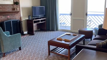A hotel room with a patterned carpet, a television on a wooden stand, a small table, a sofa, and a balcony with a view.