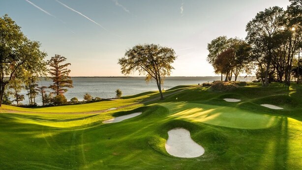Campo de golf con un cuerpo de agua al fondo, rodeado de árboles y bunkers de arena.
