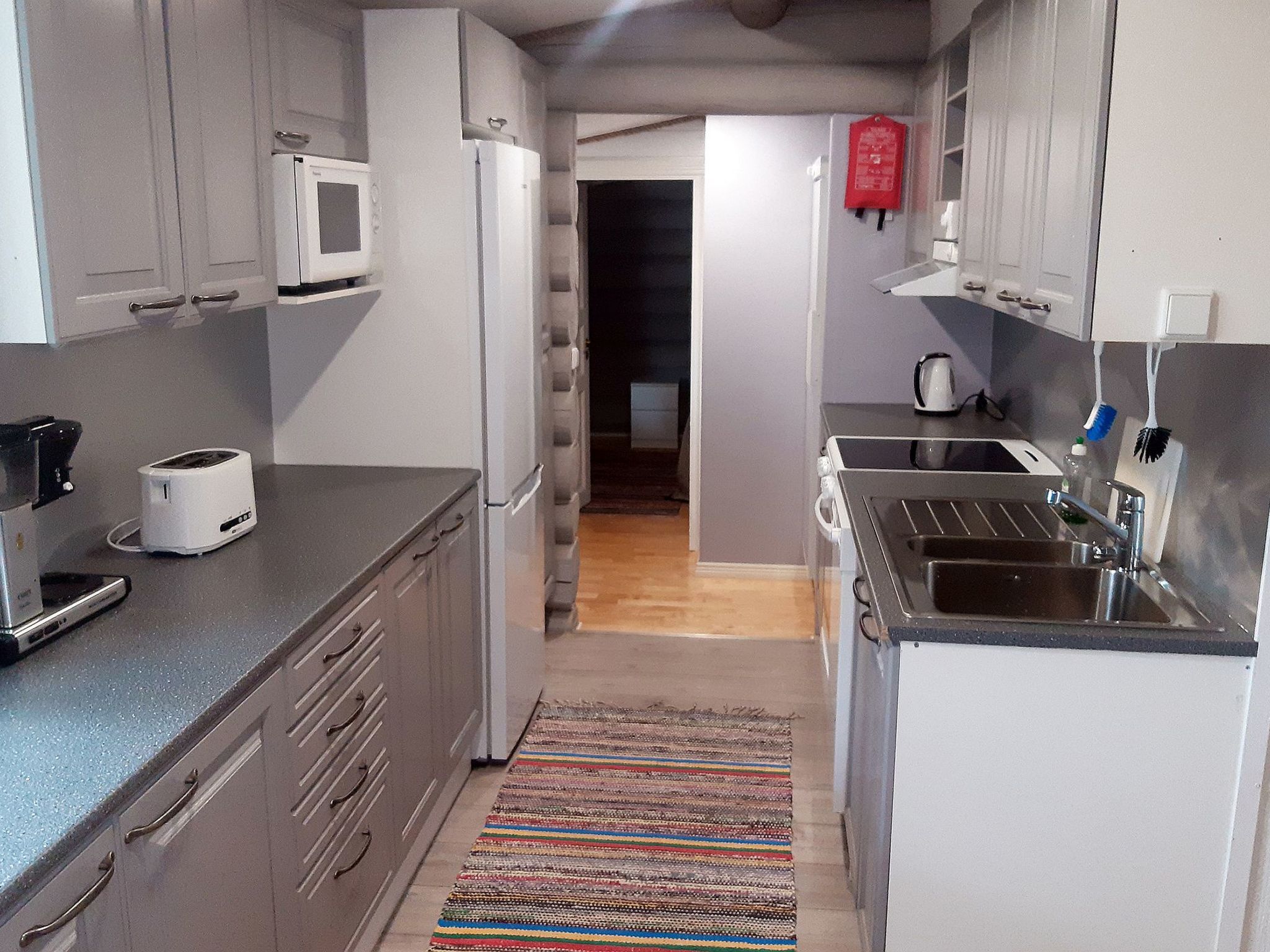 A compact kitchen with a microwave, toaster, and sink. A colorful rug is on the floor, and a red fire extinguisher is mounted on the wall.