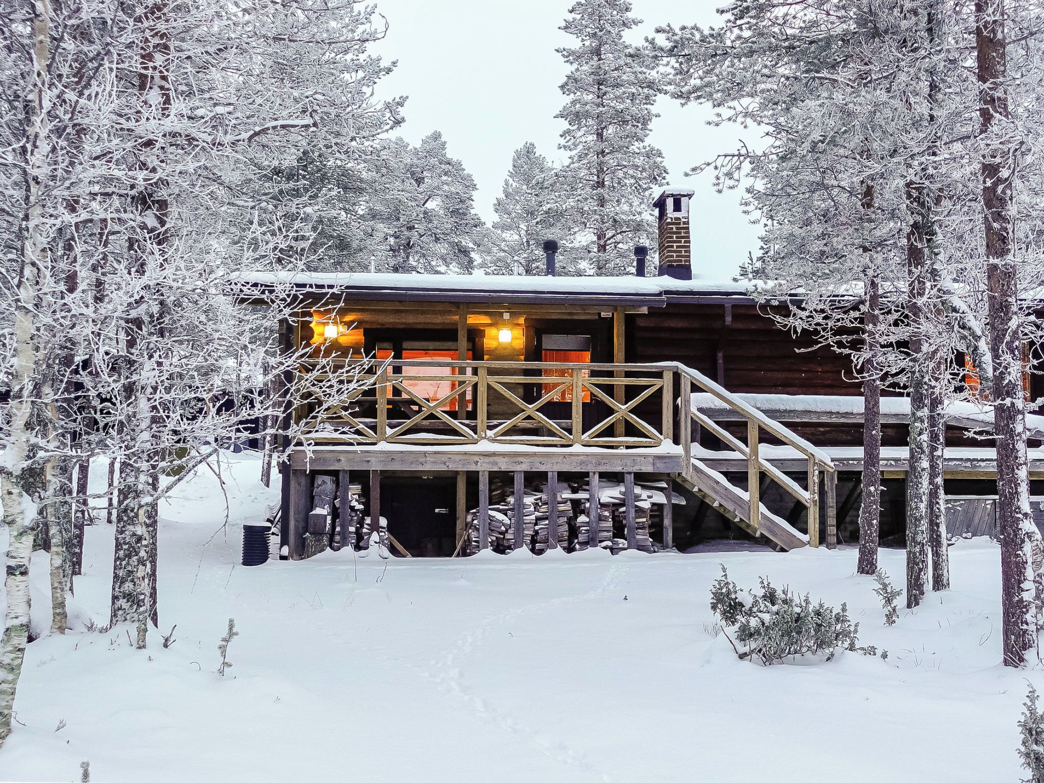 A wooden cabin with a wooden staircase leading to a deck, surrounded by snow-covered trees.