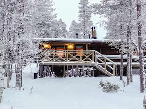 Una cabaña de madera con una escalera de madera que conduce a una terraza, rodeada de árboles cubiertos de nieve.