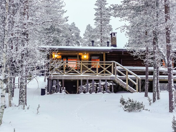 Una cabaña de madera con una escalera de madera que conduce a una terraza, rodeada de árboles cubiertos de nieve.