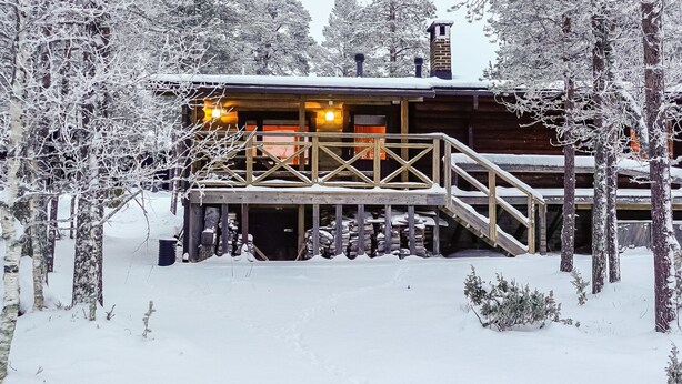 A wooden cabin with a covered deck and a chimney, surrounded by snow-covered trees.