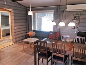 Interior de una cabaña de troncos con comedor, un sofá y una ventana con persianas.