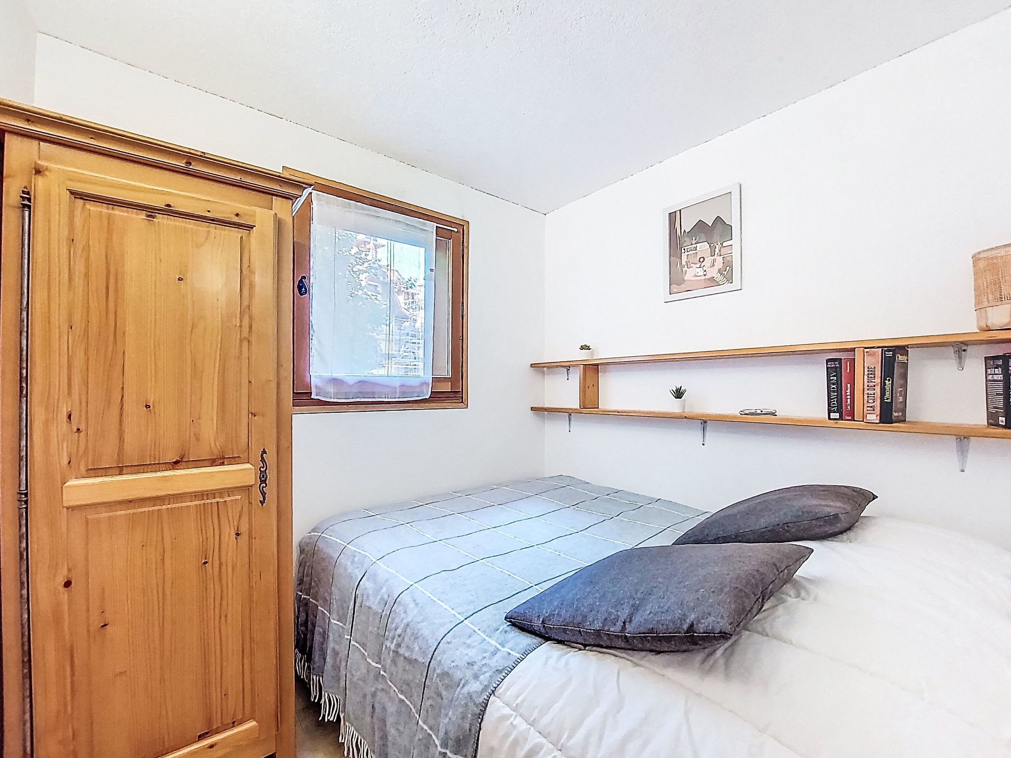 A bedroom with a wooden wardrobe, a bed with a grey and white striped blanket, two pillows, a window with a white curtain, a wall-mounted shelf with books, and a framed picture.
