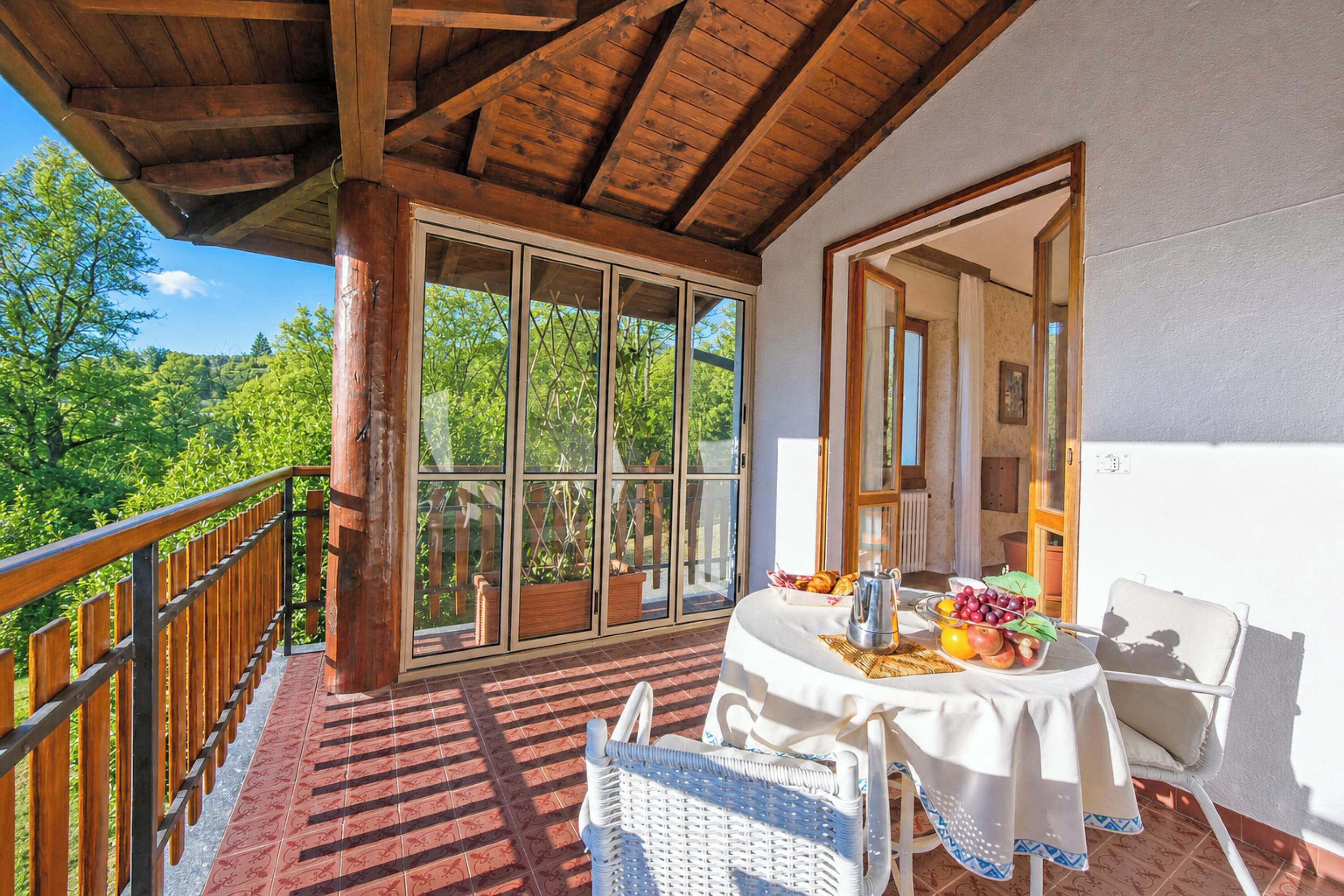 A covered balcony with a table set for dining, a wicker chair, and a view of greenery.