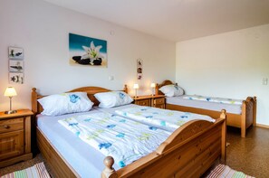 Quarto com três camas de madeira, cada uma com uma mesa de cabeceira e uma luminária. Há um quadro na parede acima das camas.