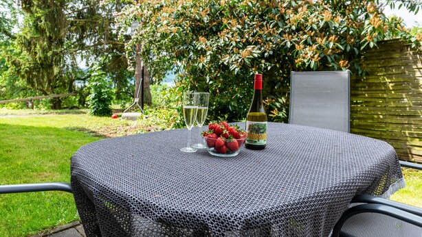 A round table with a woven tablecloth, a bowl of strawberries, two wine glasses, and a bottle of wine.