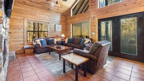 A cozy log cabin interior with a fireplace, a ceiling fan, and large windows. - Peaceful Pines: Hot Tub/Playground/Games + Eateries/Brewery/Hiking/Lake~3mi! (Broken Bow)