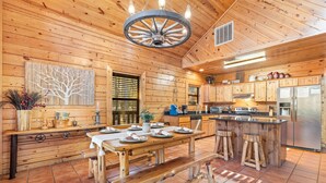 A wooden cabin interior with a dining area, kitchen, and a large wooden table with benches. - Peaceful Pines: Hot Tub/Playground/Games + Eateries/Brewery/Hiking/Lake~3mi! (Broken Bow)
