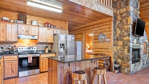 A log cabin-style kitchen with wooden cabinets, a central island with stools, a stone fireplace, and a television mounted on the stone wall. - Peaceful Pines: Hot Tub/Playground/Games + Eateries/Brewery/Hiking/Lake~3mi! (Broken Bow)