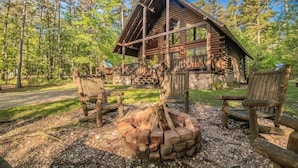 A rustic cabin with a stone foundation, wooden exterior, and a fire pit in the foreground. - Peaceful Pines: Hot Tub/Playground/Games + Eateries/Brewery/Hiking/Lake~3mi! (Broken Bow)