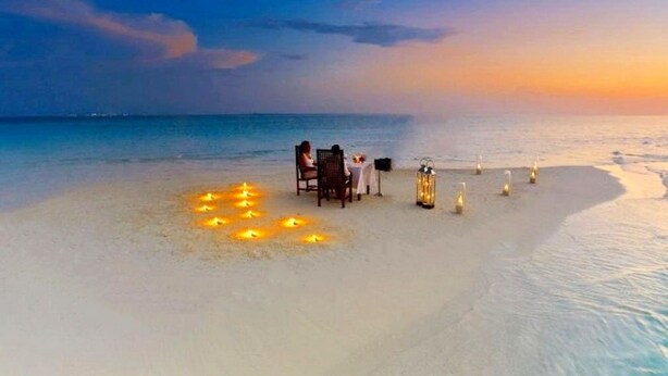 A beach setting with a table and chairs, a small table with a lamp, and candles on the sand. - Adroit Beach Inn (Guraidhoo)