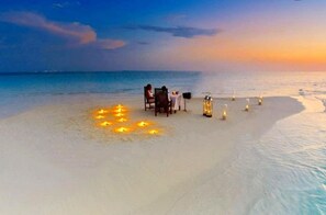 Ein Strandbild mit Tisch und Stühlen, einem kleinen Tisch mit Lampe sowie Kerzen im Sand.