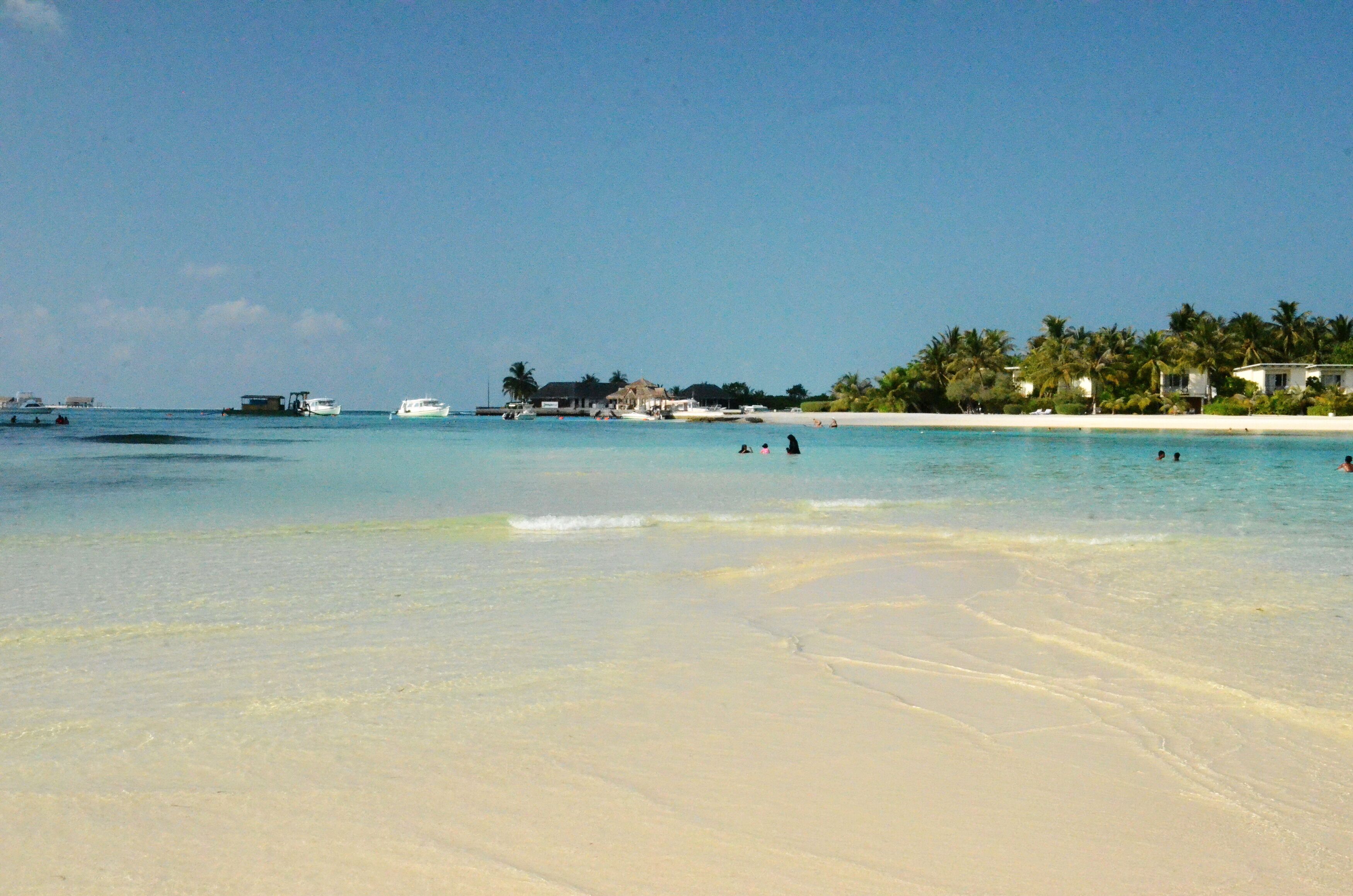 Una playa con agua turquesa cristalina, una orilla arenosa y algunos edificios al fondo.