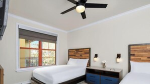A hotel room with two beds, a ceiling fan, a window with a view, and a nightstand with a lamp.
