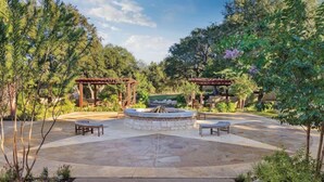 A circular stone patio with a central fire pit, surrounded by two wooden pergolas and benches.