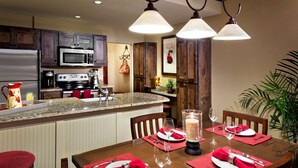 A modern kitchen with dark wooden cabinets, a stainless steel refrigerator, and a granite countertop. There is a dining area with a wooden table set for four, red napkins, and a candle centerpiece.