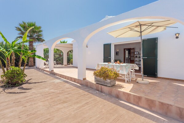 A white building with a covered patio area, a table with chairs, and a potted plant.