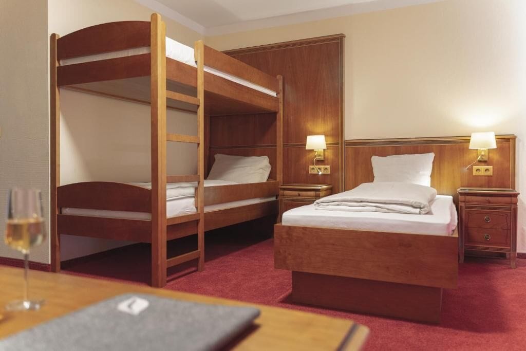 Una habitación de hotel con una cama nido, una cama individual, dos mesitas de noche con lámparas, un mesón de madera y una copa de vino sobre una mesa.