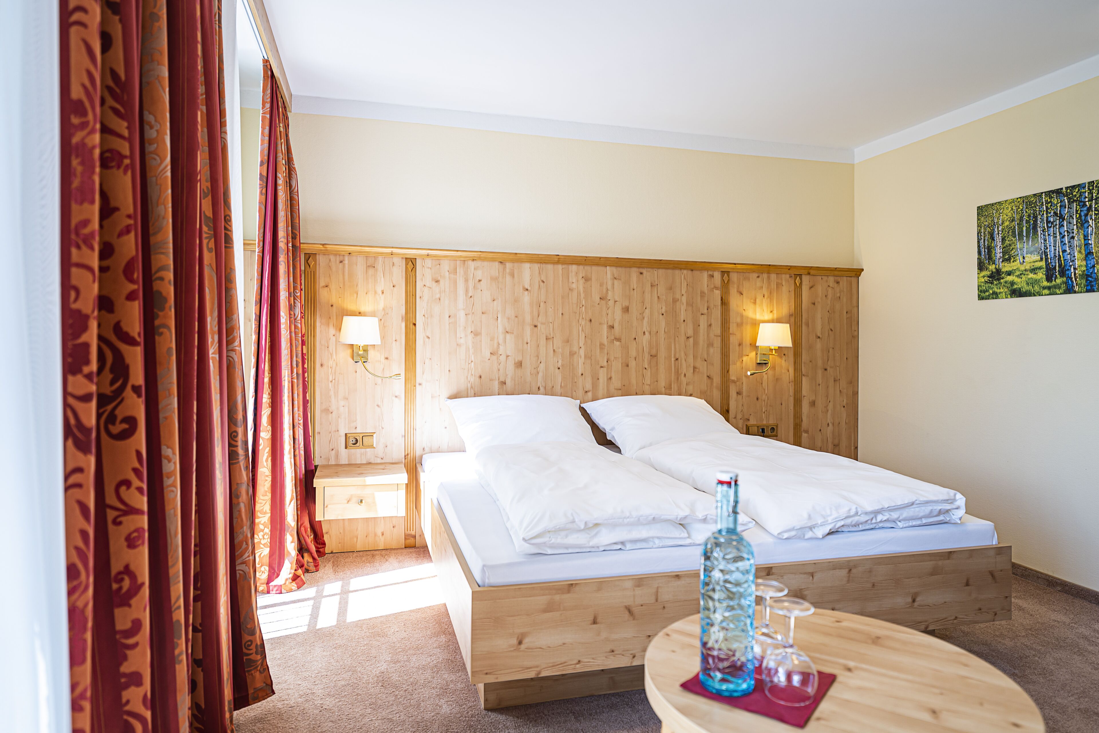 Ett hotellrum med en säng i trä, två sänglampor, ett litet bord med en flaska och glas, och en tavla på väggen.